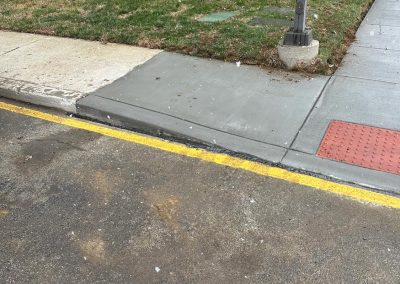 Sidewalk corner with stone building, bushes, and a snowy ground. Yellow curb line visible.