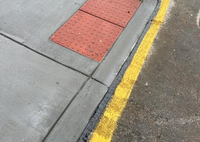 Sidewalk corner with tactile paving and yellow-painted curb, adjacent to a road.