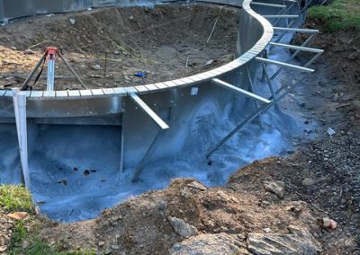 Partially constructed in-ground pool with metal frame surrounded by dirt and trees.