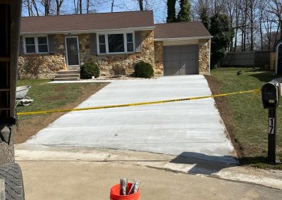Freshly poured concrete driveway of a suburban house, with yellow caution tape and a red bucket in front.