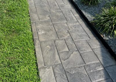 Gray stone walkway bordered by green grass and trimmed plants on a sunny day.
