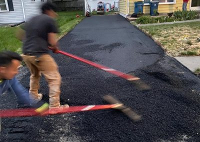 Workers paving a driveway with asphalt outside a house on a residential street.