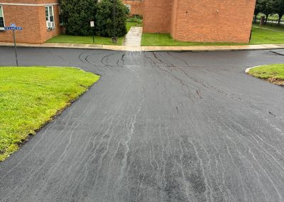 Freshly paved asphalt road with tire marks, bordered by green grass, leading to brick buildings on an overcast day.