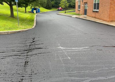 Freshly paved driveway beside a brick building, with trees and a parked red car in the background.