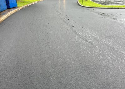 Red truck parked on a wet, freshly paved road near blue dumpsters and surrounded by green trees.