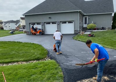 Workers paving a driveway in front of a gray house with a garage and machinery.