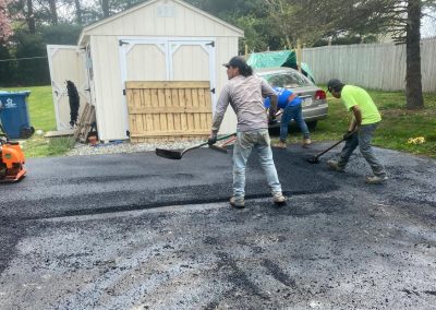 Workers laying asphalt in front of a shed and vehicle in a yard.