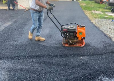 A worker uses a vibrating plate compactor on a driveway, smoothing fresh black asphalt. Another worker is in the background.