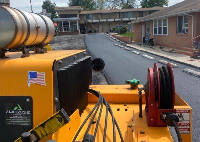View from a road paver machine laying fresh asphalt in a residential area, with houses and trees in the background.