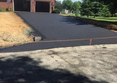 Freshly paved driveway leading to a brick house with a double garage, surrounded by a grassy lawn.