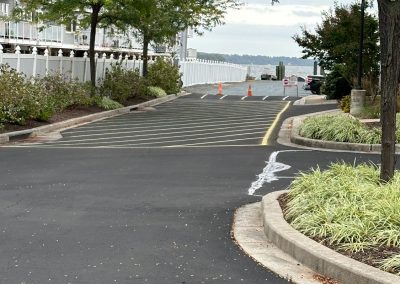 Paved road with speed bumps, bordered by trees and shrubs. Orange cones in the distance near a white fence.