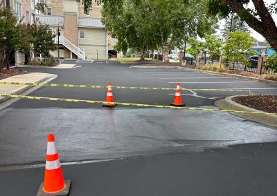 Freshly paved road with orange cones and caution tape blocking access, surrounded by trees and buildings.