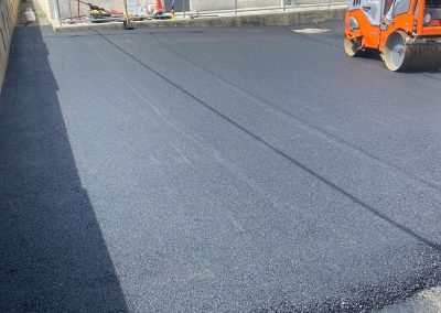 Freshly paved asphalt parking lot with an orange roller compactor. Building and sky in the background.