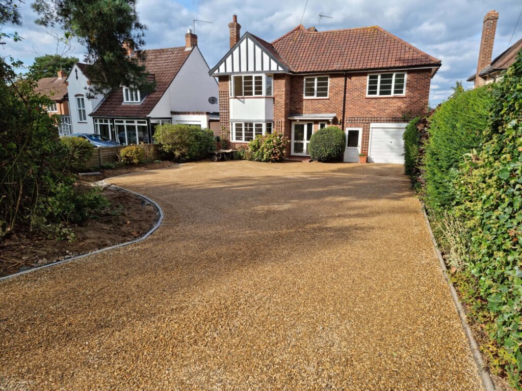 House with a gravel driveway, surrounded by shrubs and trees, adjacent to two-story homes under a partly cloudy sky.