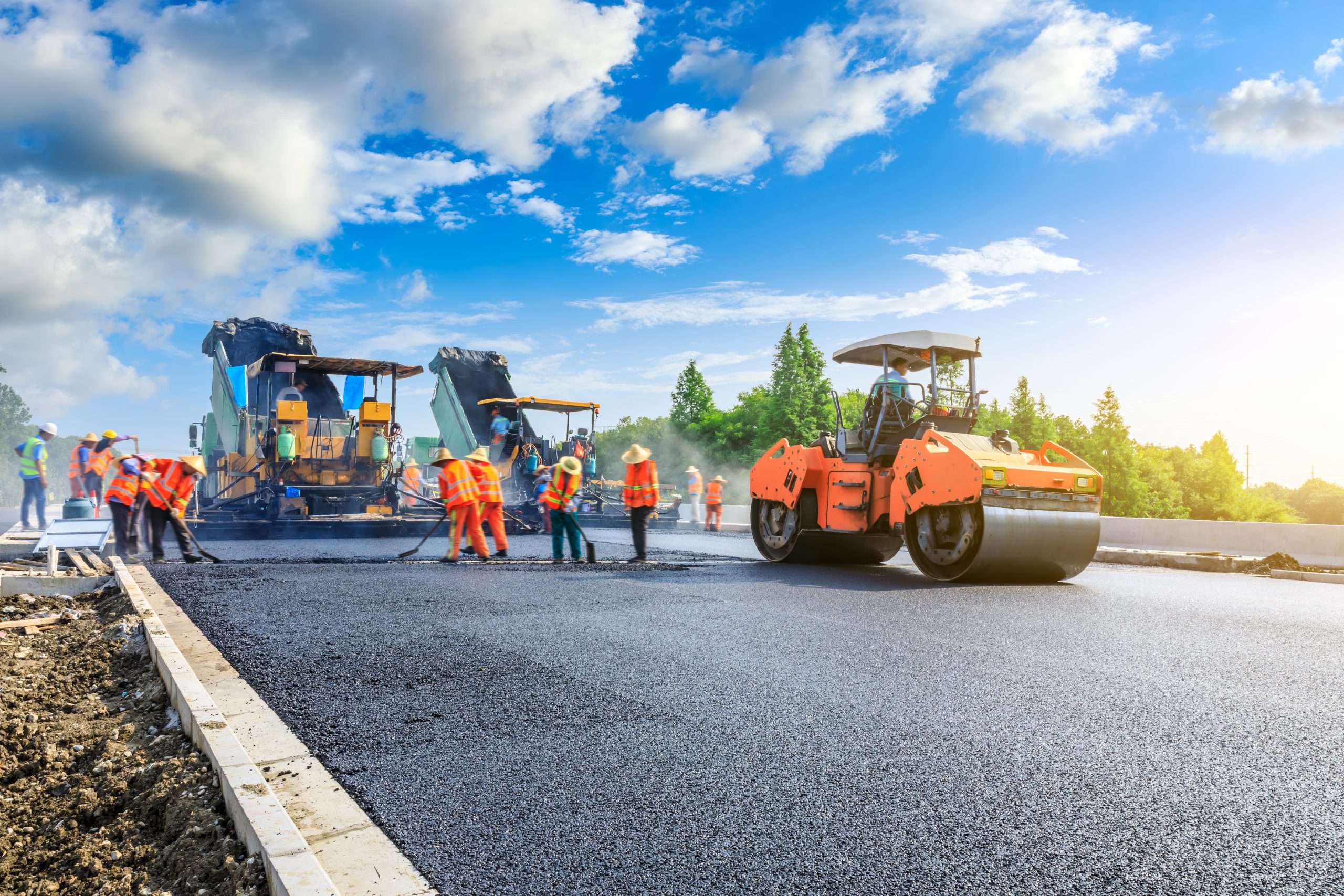 Under a blue sky with clouds, professional asphalt paving is underway as workers and machinery meticulously craft the road's smooth surface.