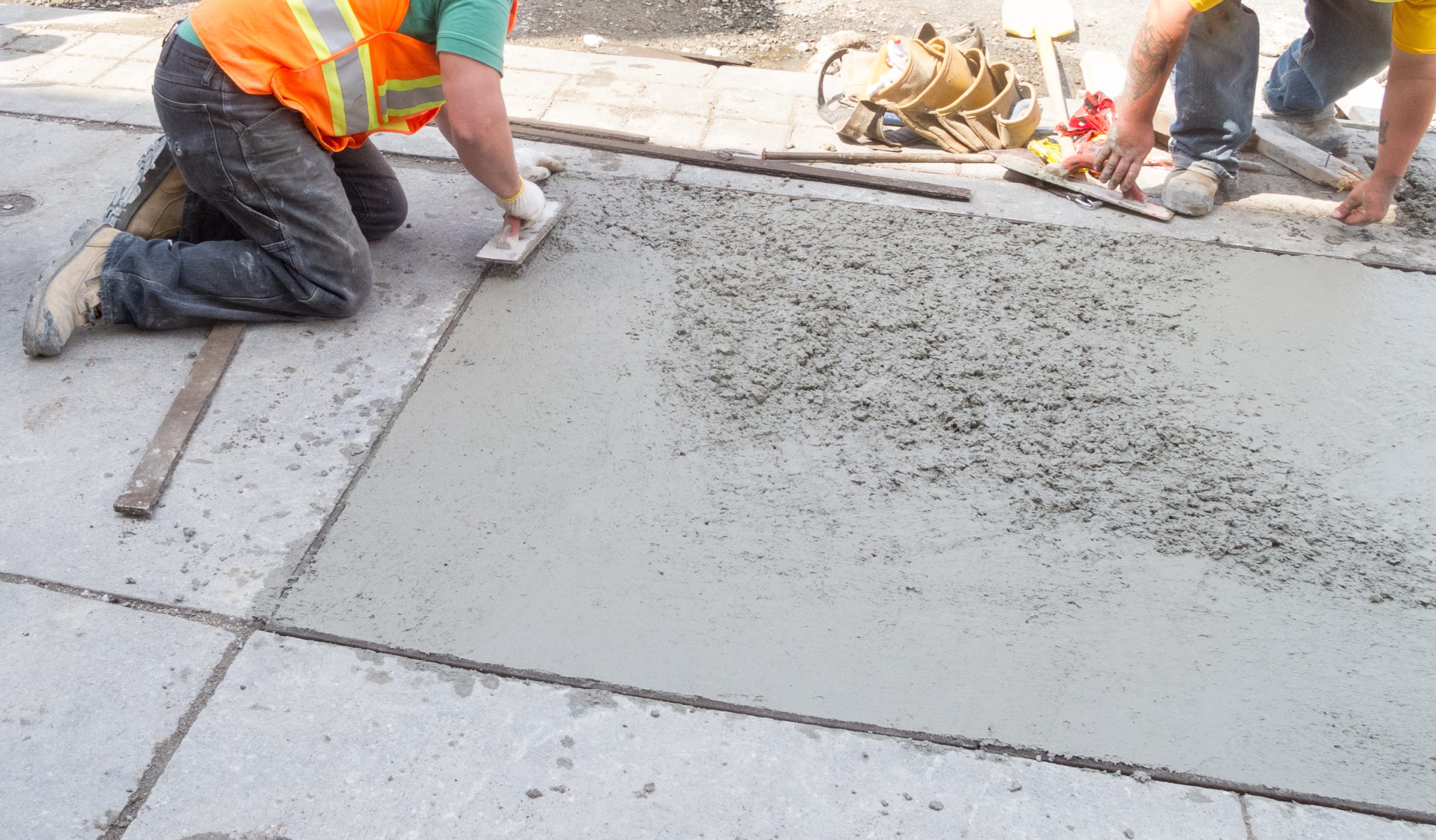 Workers in safety vests smoothing wet concrete on a section of sidewalk.