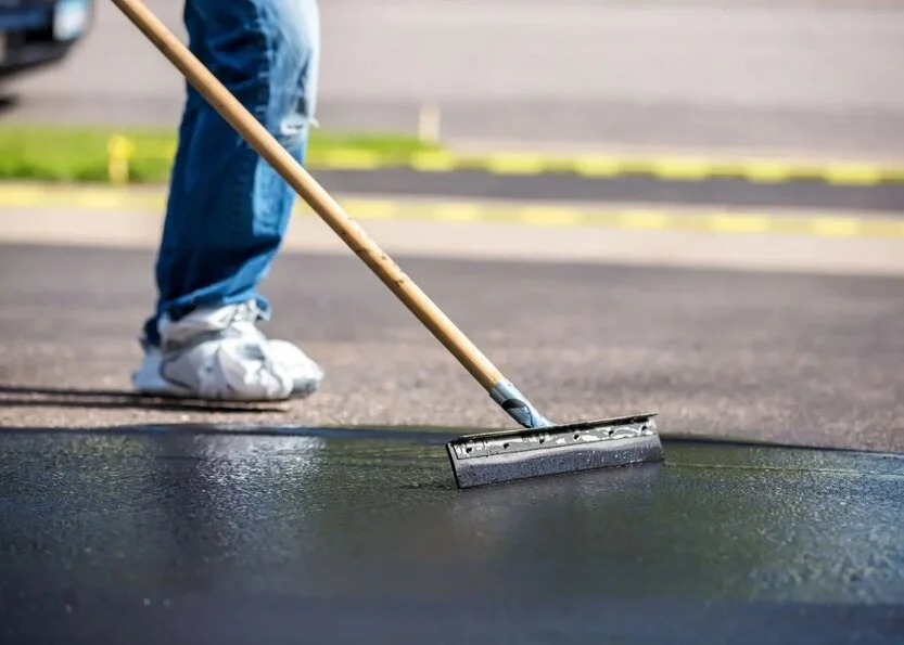 sdfsdfsd Person spreading sealant on asphalt with a long-handled squeegee, wearing jeans and white sneakers.