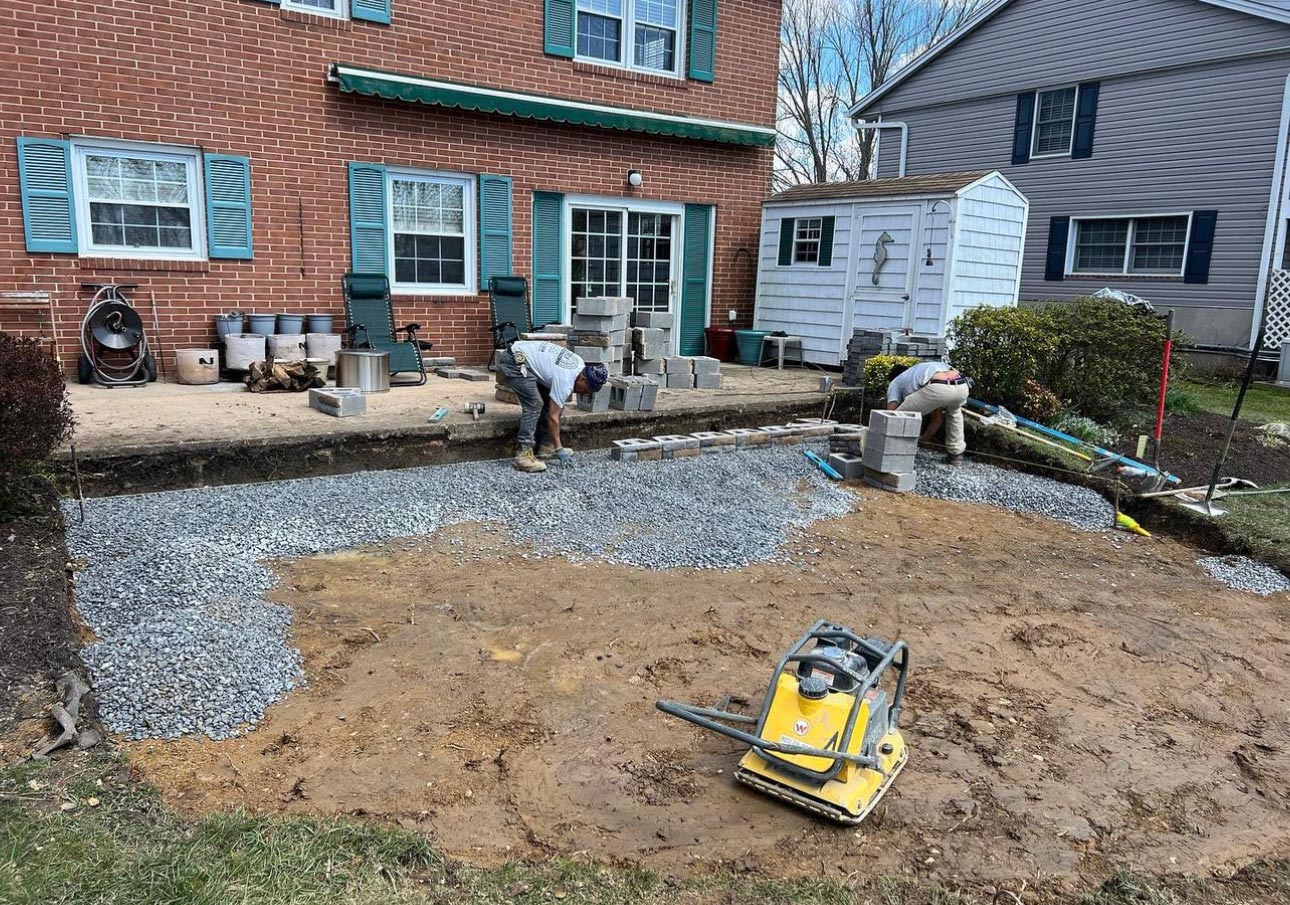 Two workers build a patio with gravel and concrete blocks in a backyard outside a brick house.